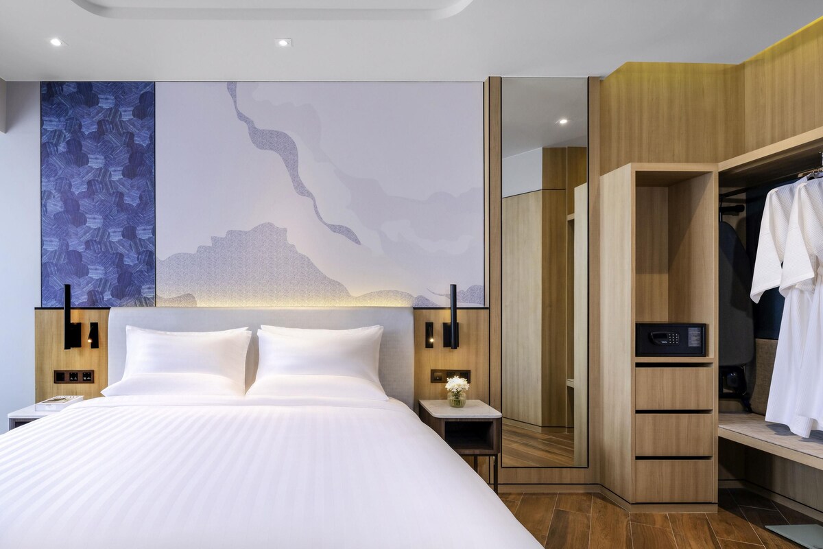 A modern hotel room with a large bed, wooden wardrobe, and a wall with abstract art.