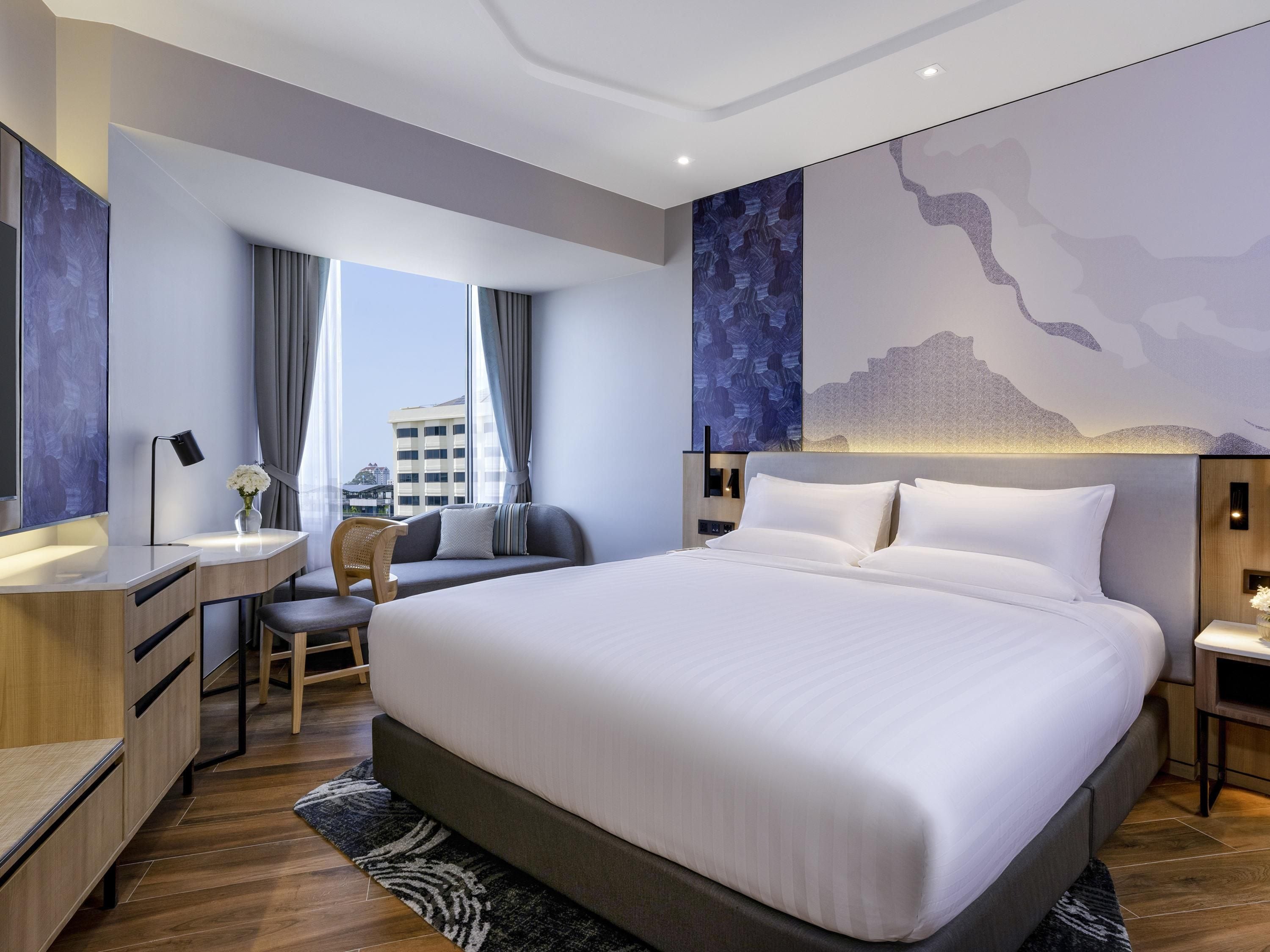 Ein modernes Hotelzimmer mit einem großen Bett, einem Schreibtisch, einem Sessel und Blick auf die Stadt.