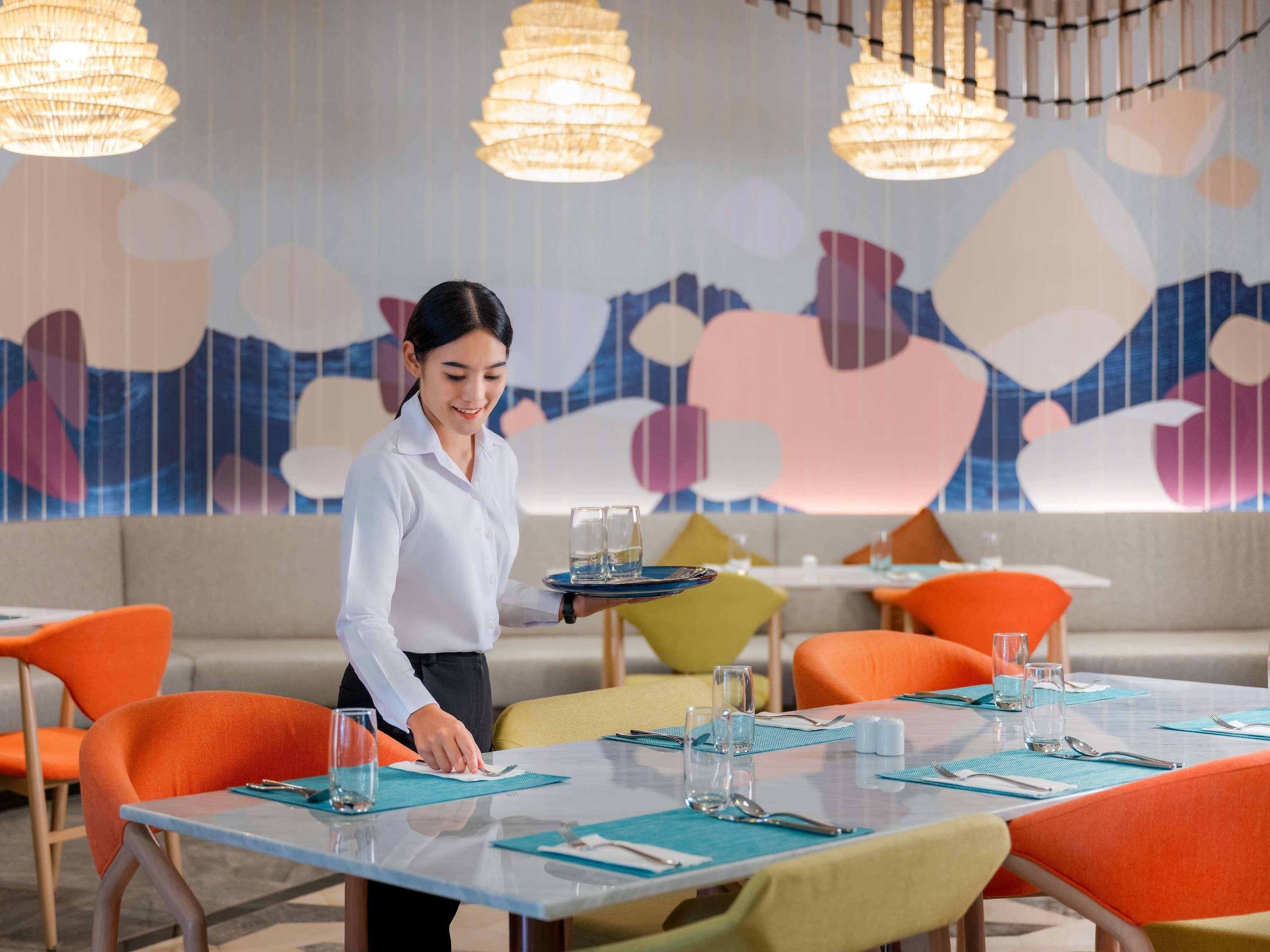A modern dining area with a waiter serving a tray of glasses. Tables are set with blue placemats, and the room features colorful abstract wall art.