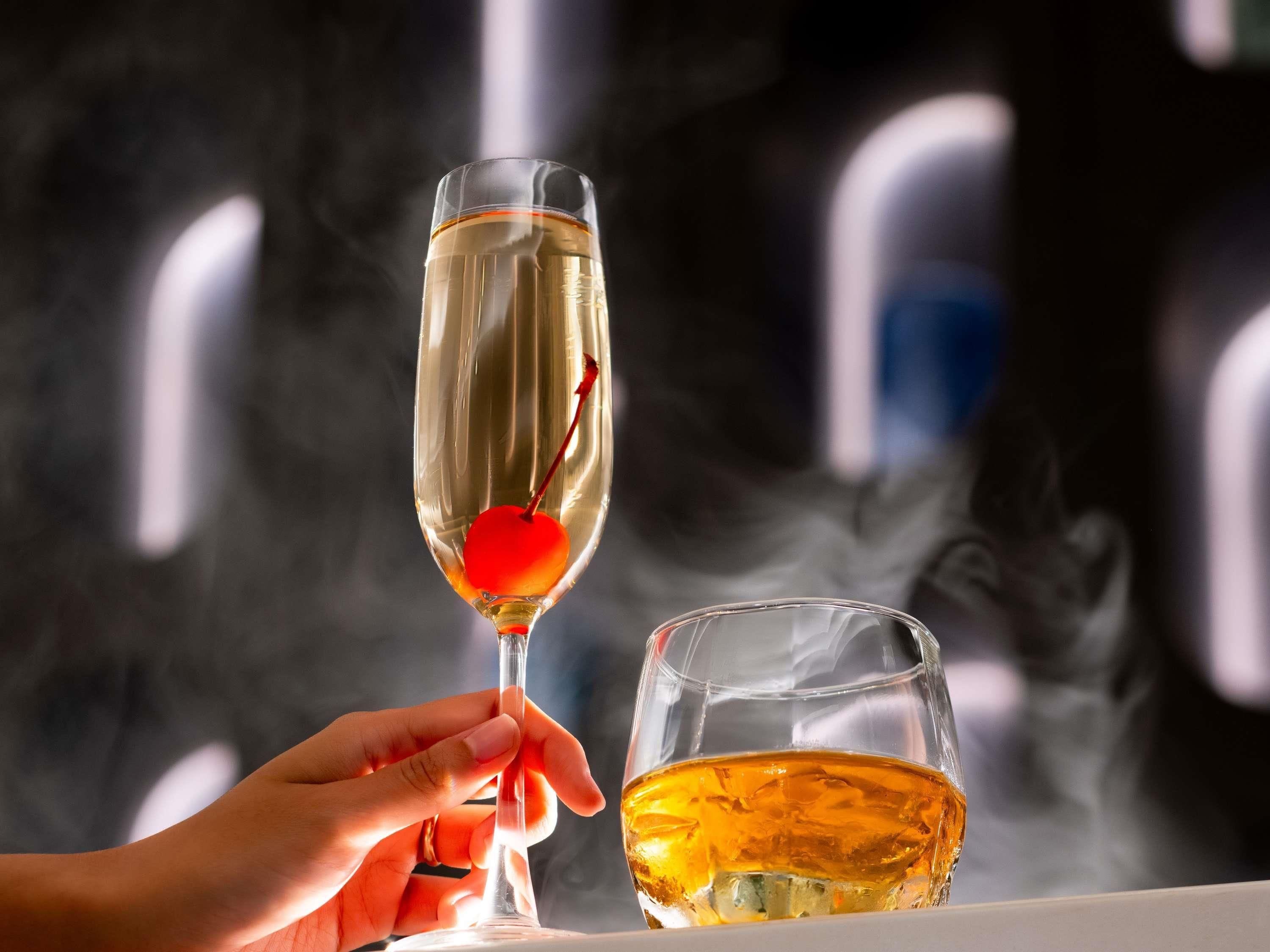 Une main tenant une coupe à champagne contenant un objet rouge en forme de cœur, à côté d’un verre rempli d’un liquide doré.