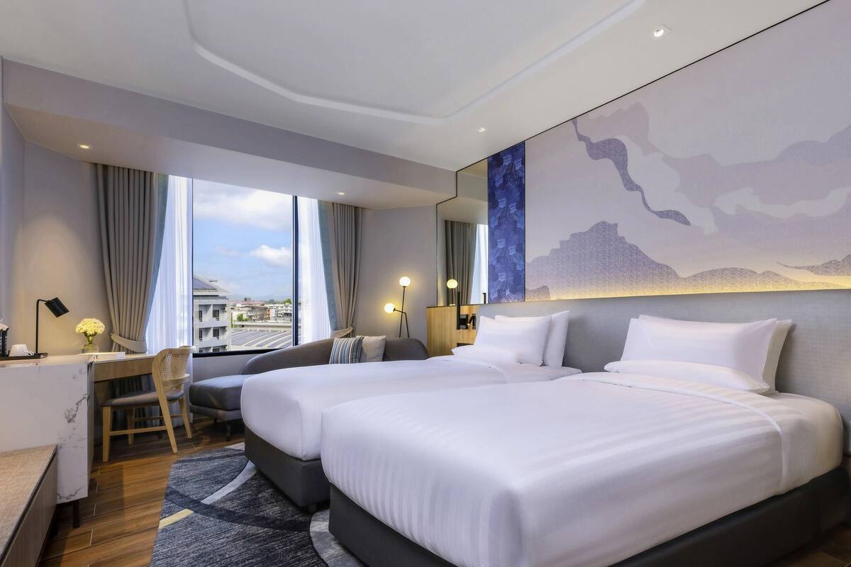 A hotel room with two beds, a desk, a chair, a large window with a city view, and a modern wall art piece.