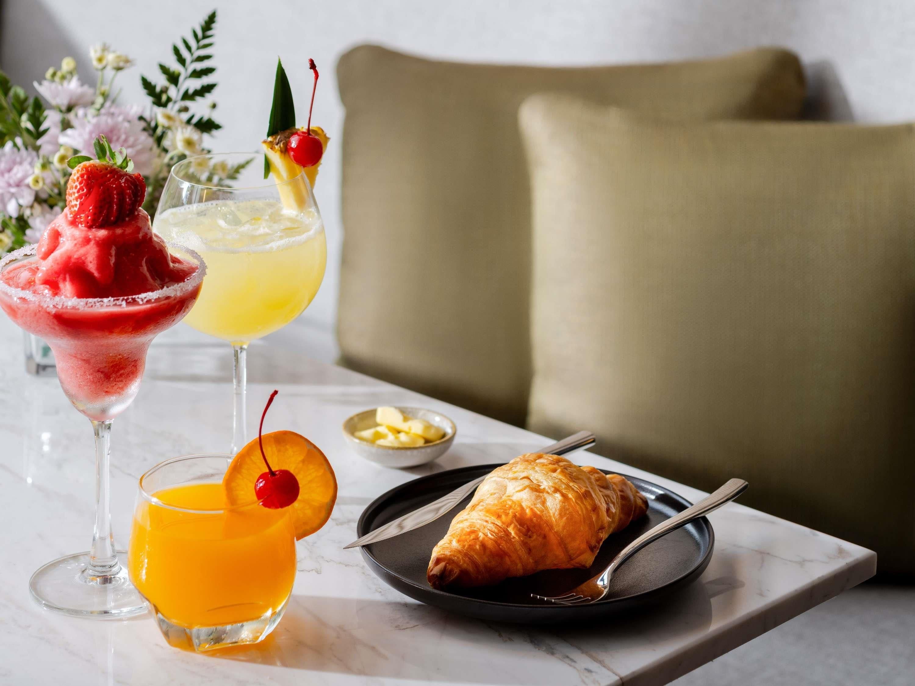 Ein Tisch mit einem Dessertgetränk, einem Fruchtsaft und einem Croissant auf einem Teller.