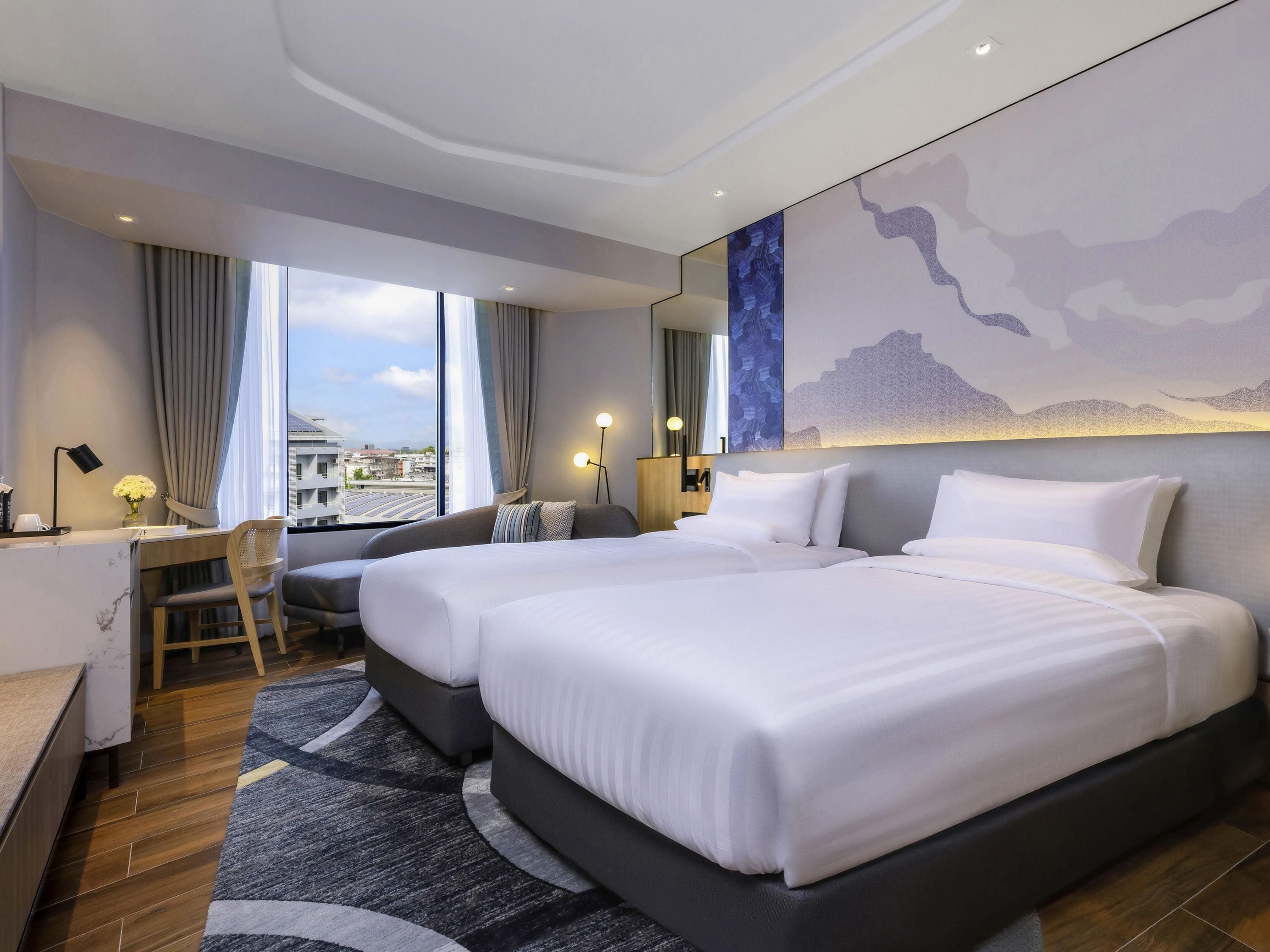 Una camera d'albergo con due letti, una scrivania, una sedia, un'ampia finestra con vista sulla città e un'opera d'arte moderna appesa al muro.