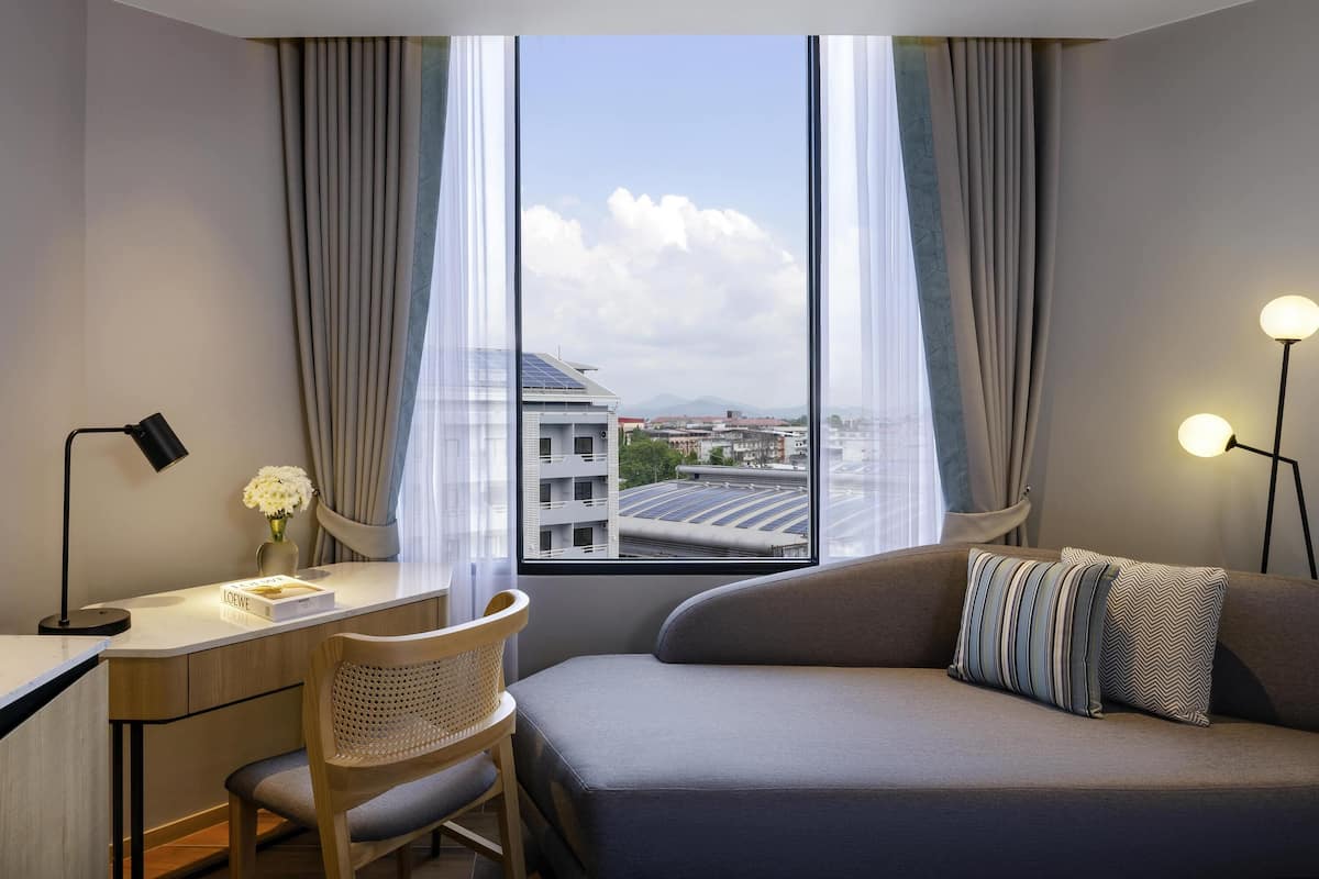 Una habitación de hotel moderna con escritorio, silla y sofá, ventanas amplias con cortinas, y vista a edificios y paneles solares.