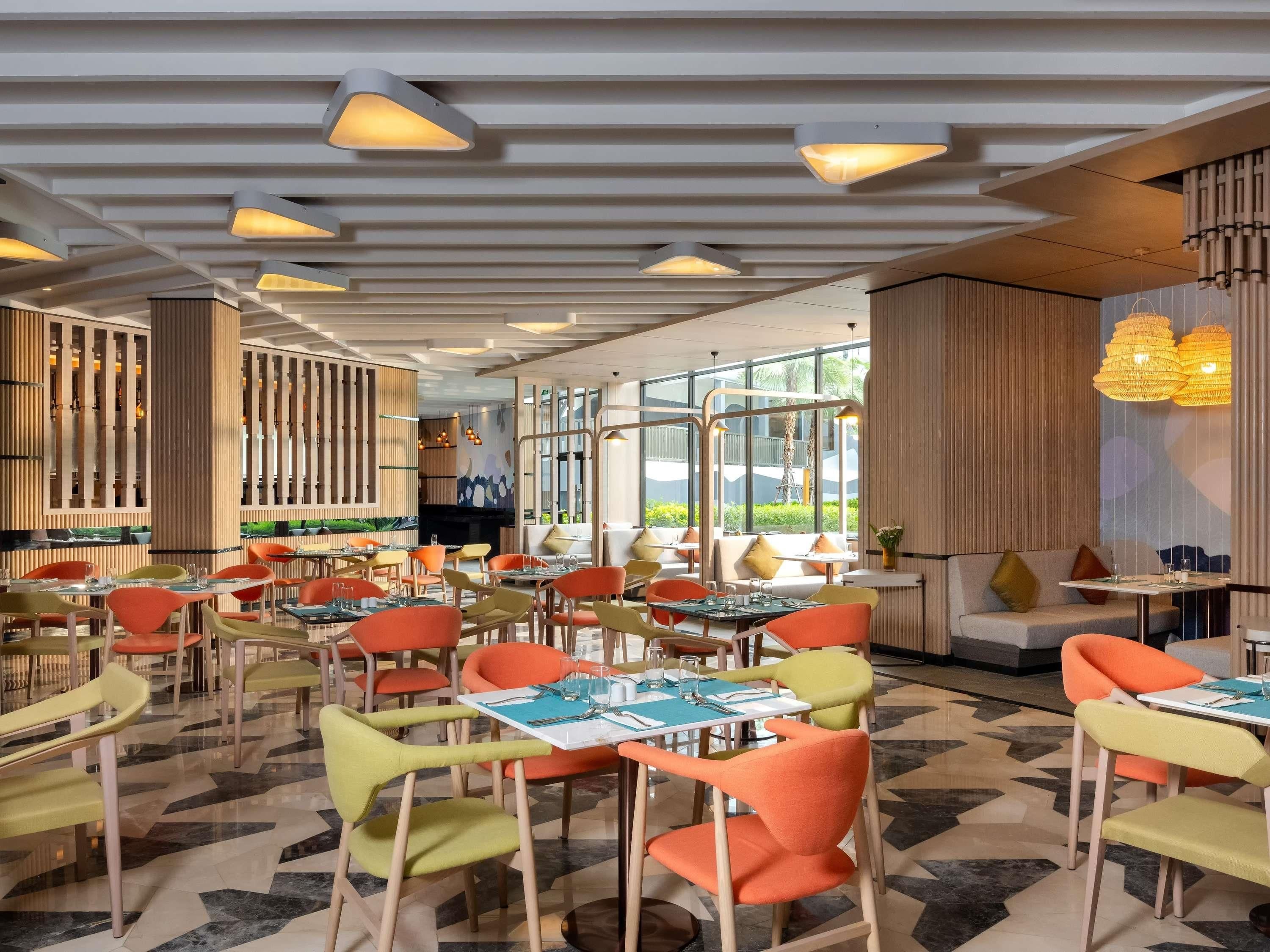 A modern restaurant interior with multiple dining tables and colorful chairs, large windows, and contemporary lighting fixtures.