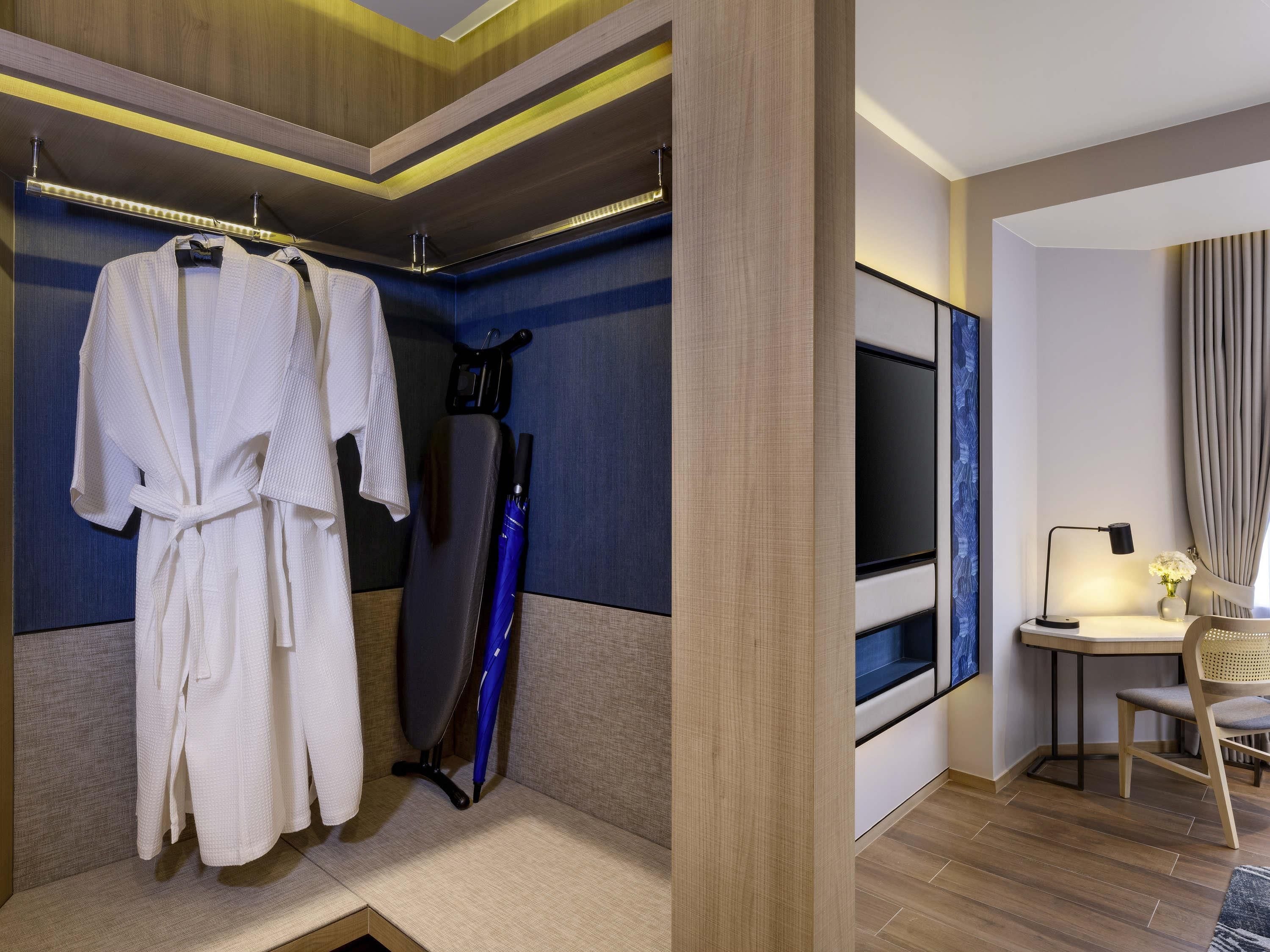 Habitación de hotel con perchero, un paraguas azul, escritorio con lámpara y un espejo grande.