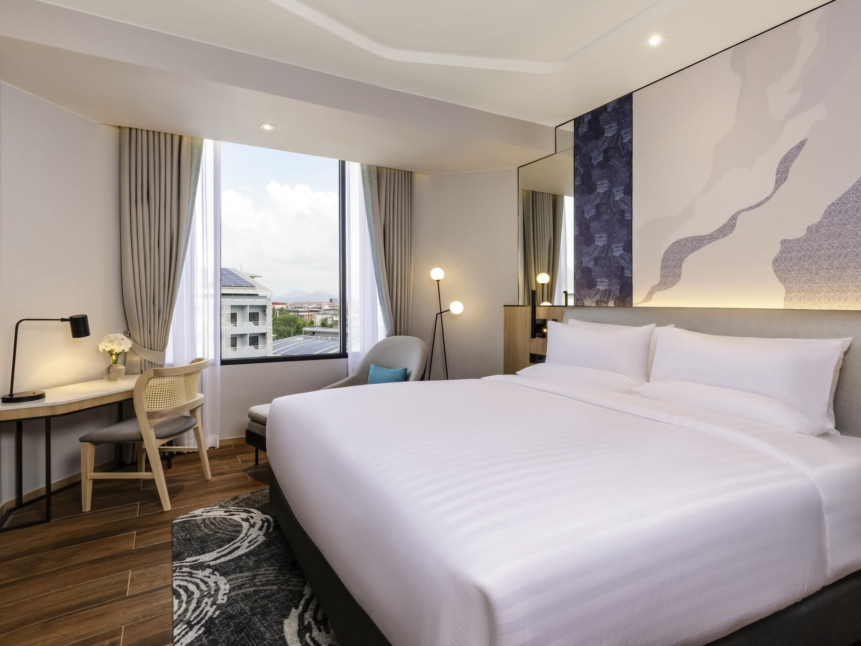 Ein modernes Hotelzimmer mit einem großen Bett, einem Schreibtisch mit Stuhl, einer Stehlampe und Blick auf die Stadt durch das Fenster.