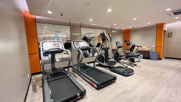 Un gimnasio con cintas de correr, elípticas y una pequeña mesa con un bote de basura.