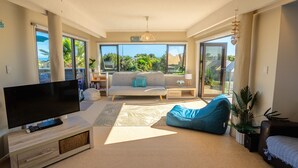 A modern living room with a flat-screen TV, a sofa, a coffee table, and a large window offering a view of greenery. - Pohara Beach Escape - Pohara Beach Holiday Home (Pohara Beach)