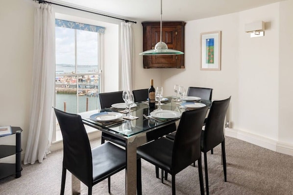 A dining room with a glass table, black chairs, a hanging light, a view of the sea, and a wooden cabinet.