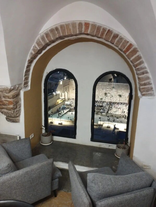 A room with two arched windows overlooking a plaza with a stone wall and a pool. There are two gray sofas and a small table with a potted plant.