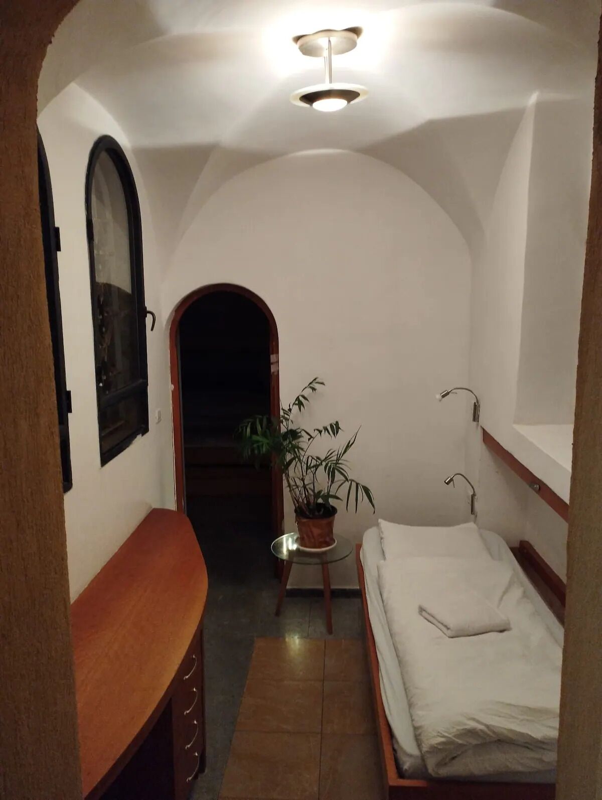 A narrow room with a bed, a small table with a plant, and a bench.