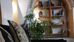 Un salon avec un canapé noir, un coussin décoratif, une plante en pot et une bibliothèque remplie de livres et d’objets décoratifs.