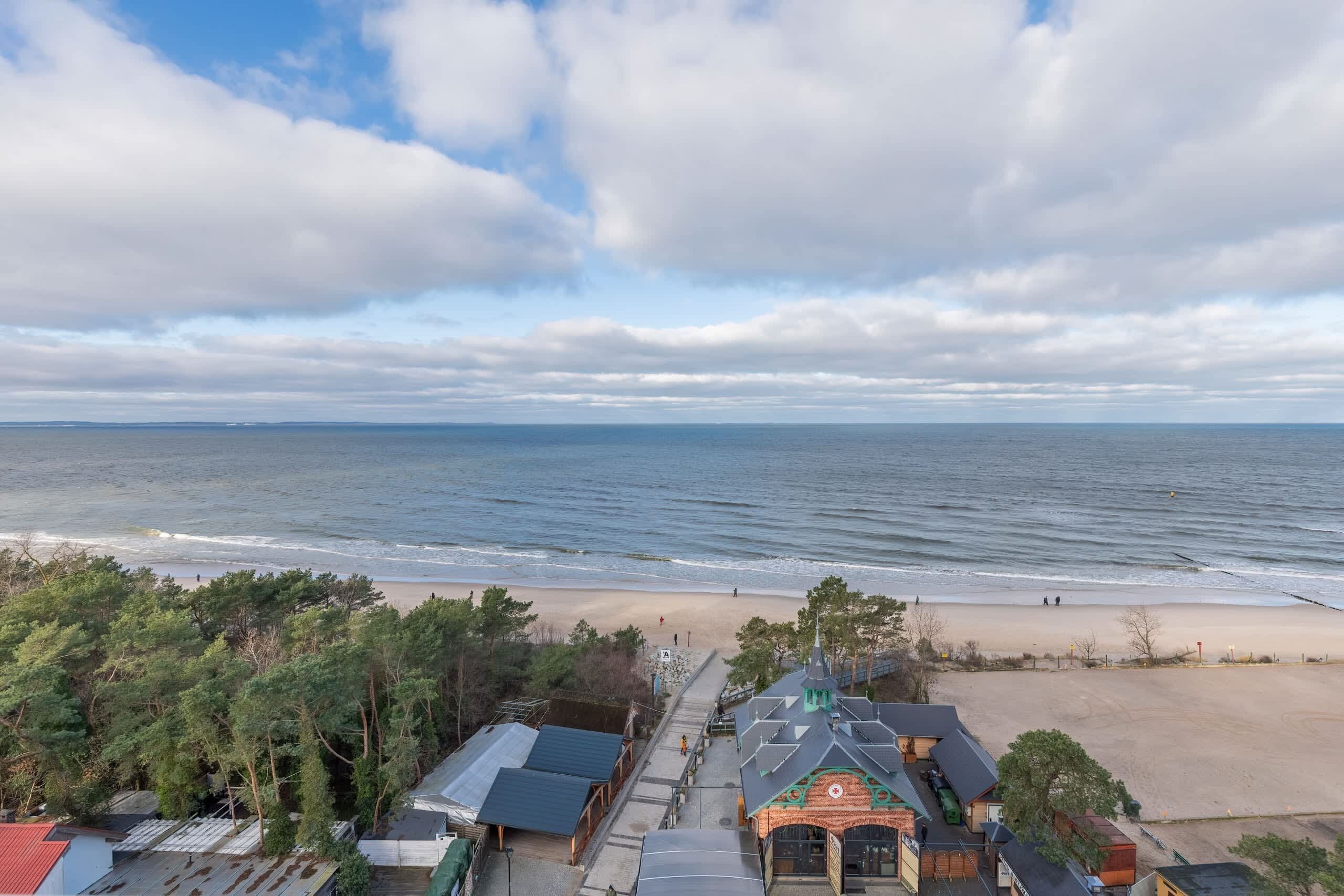 Blick auf die Küste mit Strand, einem Weg, der zu einem Gebäude mit grüner Dachterrasse führt, und einem wolkenlosen Himmel.