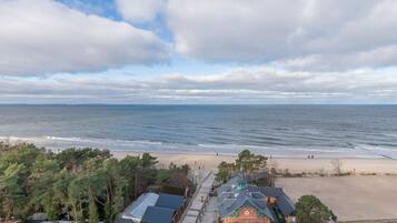 Blick auf die Küste mit Strand, einem Weg, der zu einem Gebäude mit grüner Dachterrasse führt, und einem wolkenlosen Himmel.