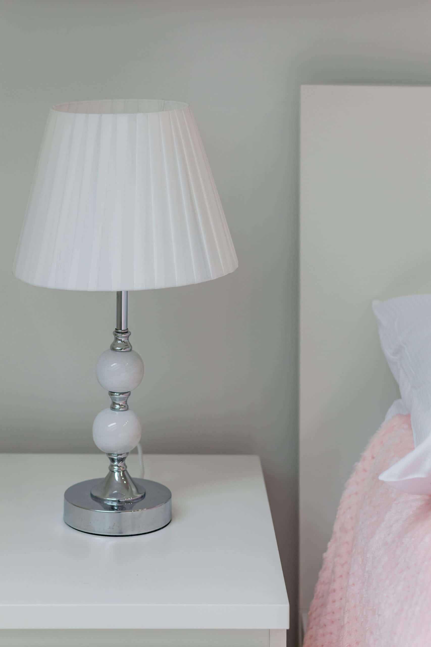 A white bedside table with a white lamp featuring a pleated shade and a metallic base.