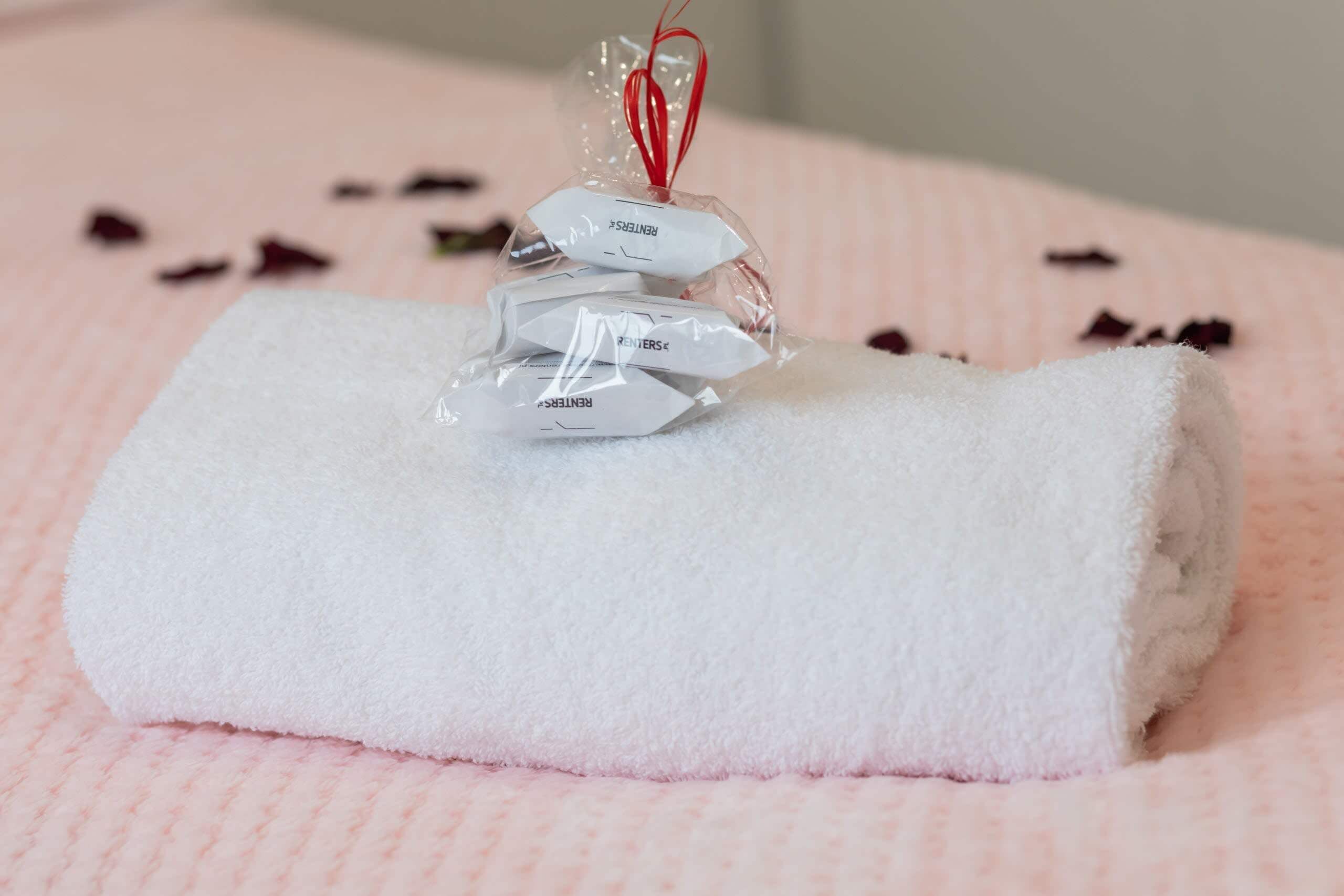 Une serviette blanche, soigneusement pliée, est posée sur une surface rose texturée, à côté d’un petit sac en plastique contenant un fil rouge et une étiquette blanche portant l’inscription « LOCATAIRES ».