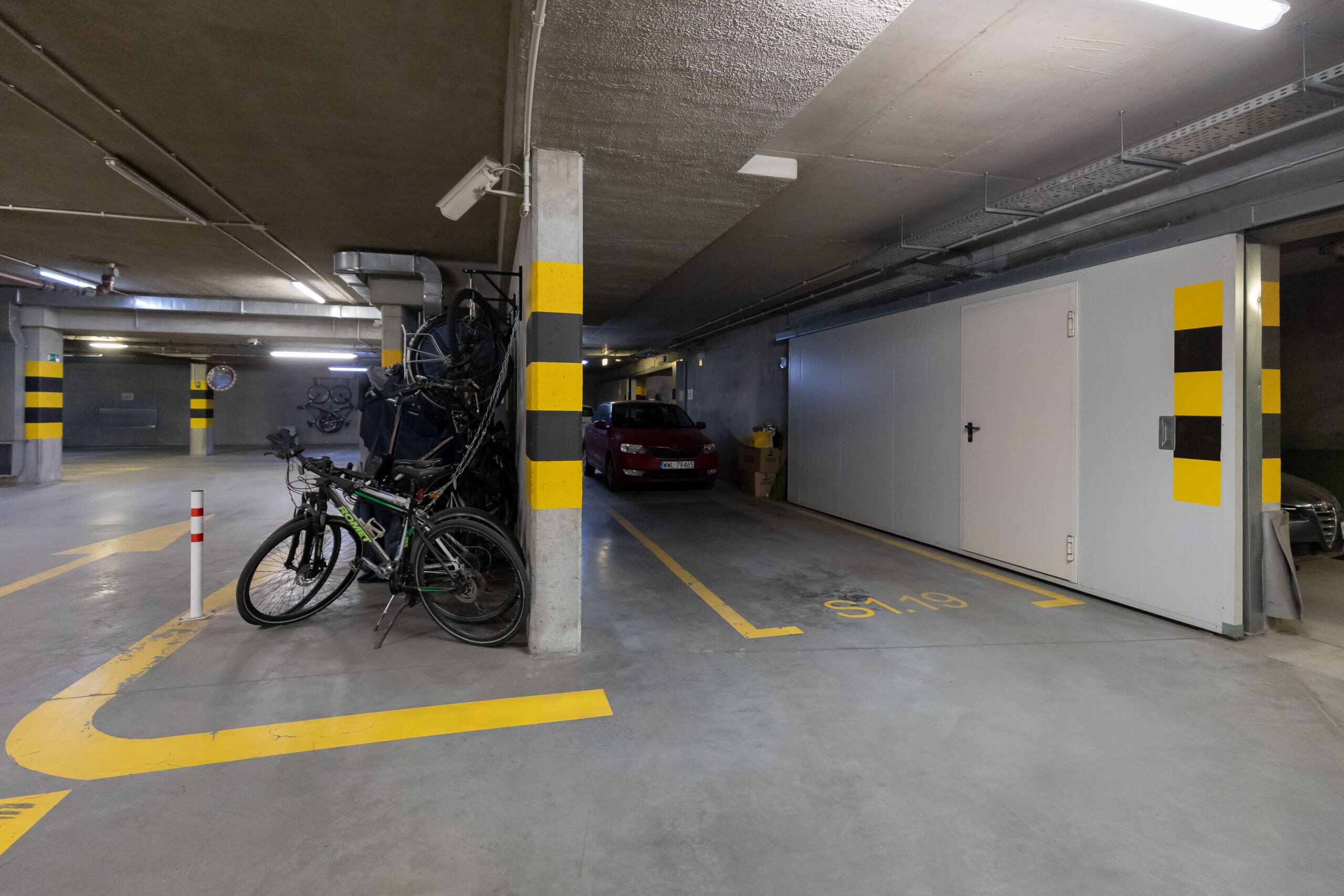 Un parcheggio sotterraneo con biciclette appoggiate a un pilastro, un'auto rossa sullo sfondo e linee direzionali gialle sul pavimento.