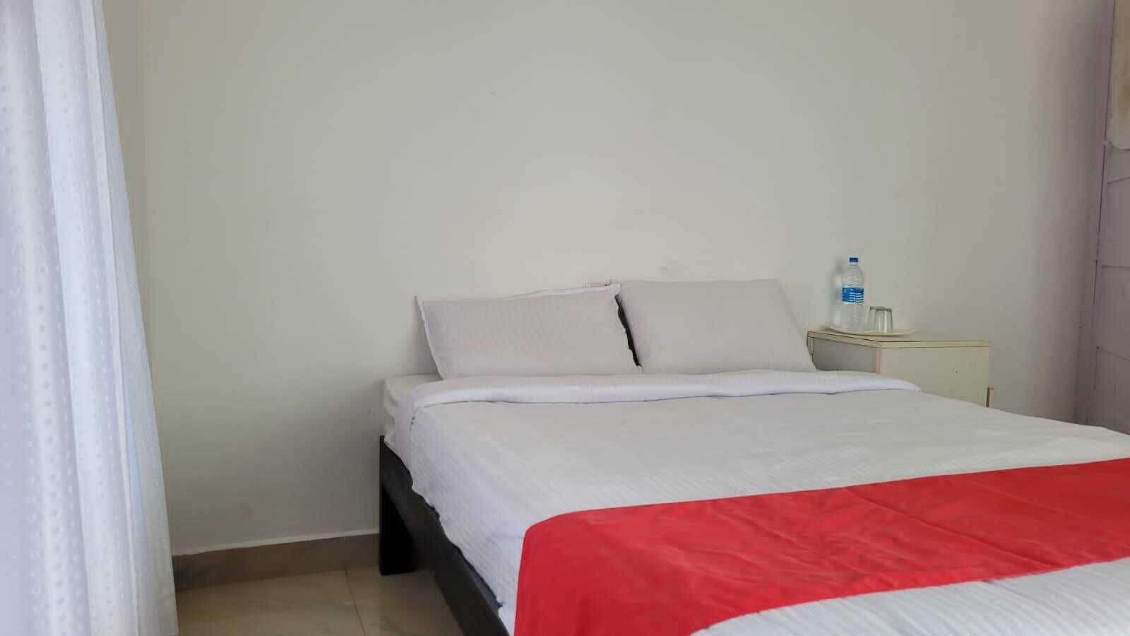 A single bed with white bedding and a red accent, a bedside table with a water bottle and glasses, and a plain white wall.