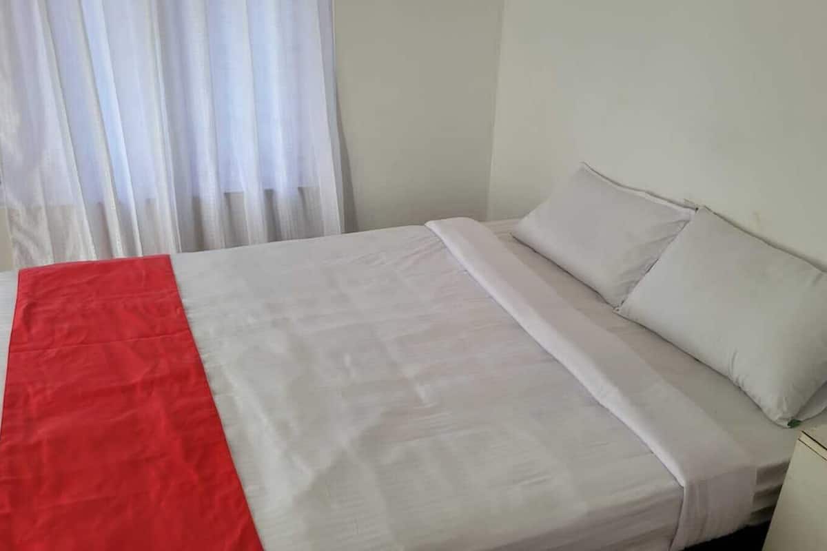 A bed with white bedding and a red accent, a white nightstand, and a window with sheer curtains.