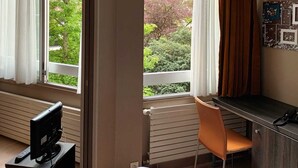 Ein Hotelzimmer mit Bett, Schreibtisch, Sessel und Fernseher, mit Blick auf Grünflächen.