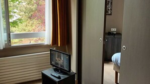 Ein Zimmer mit großem Fenster, einem Fernseher auf einem Schrank und einem Bett im Hintergrund.