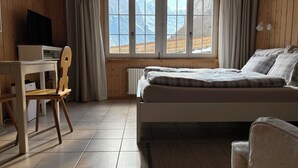 Une chambre à coucher avec un lit, un bureau et une chaise. Il y a une fenêtre d’où l’on peut admirer la vue sur les montagnes.