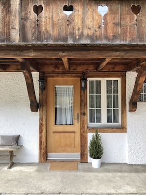 A rustic wooden building with a wooden door, a window with curtains, a small potted plant, and a bench outside.