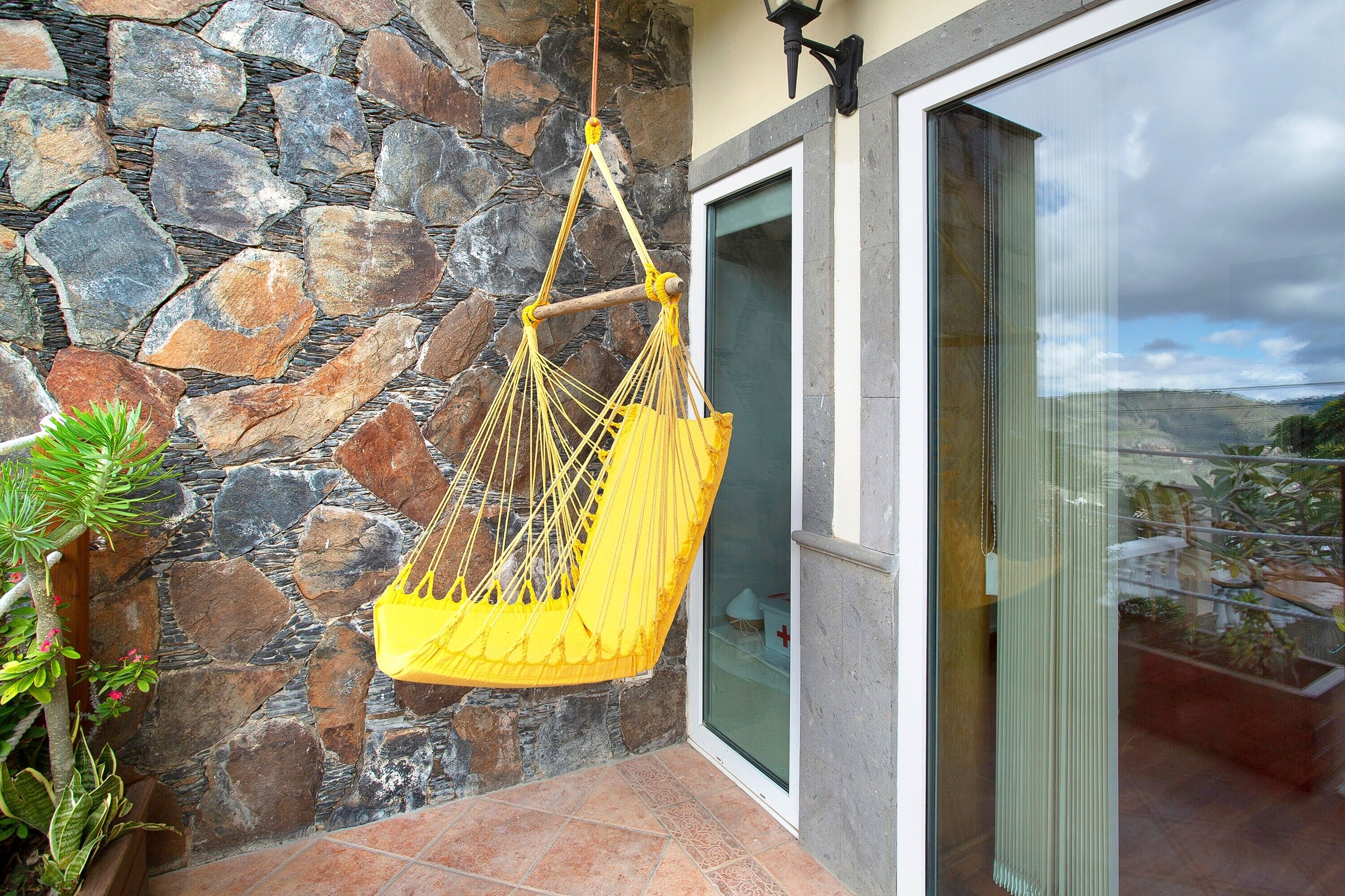 Un mur de pierre, une chaise suspendue jaune et une porte vitrée donnant sur un beau paysage.
