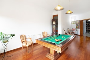 Une table de billard dans une pièce au plancher en bois, avec deux lampes suspendues et une grande horloge.