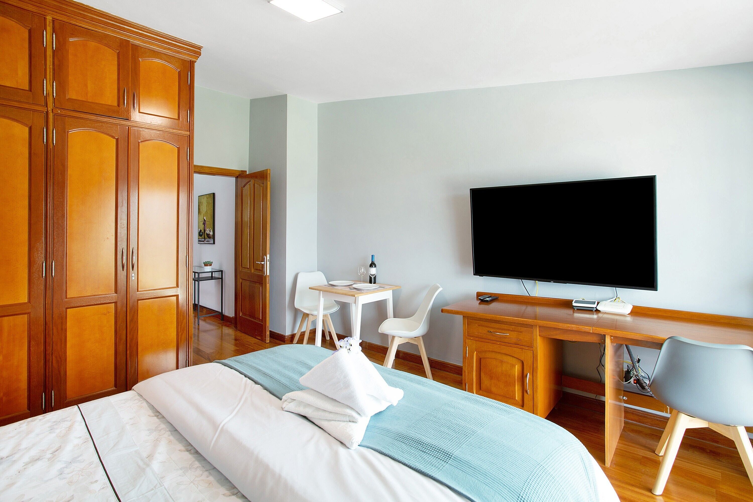 A hotel room with a bed, wooden wardrobe, desk with a flat-screen TV, two chairs, and a small table with a wine bottle.