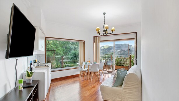 A modern living room with a flat-screen TV, a dining area with a table and chairs, and large windows offering a view of greenery and hills. - Holiday Apartment "Carmelo Y Carmela 2" with Mountain View, Private Terrace & Wi-Fi (Teror)