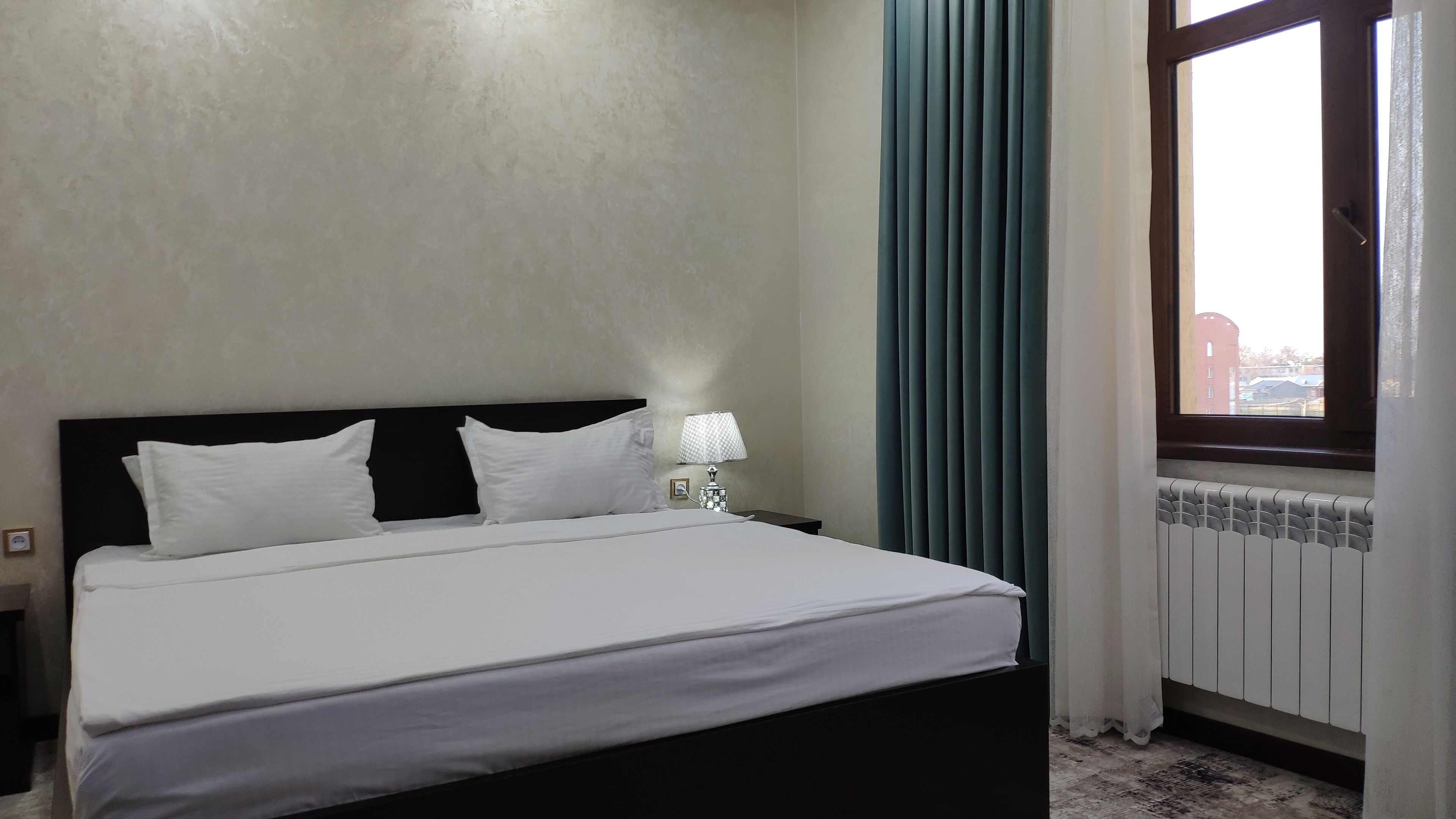 Una habitación de hotel con una cama, una mesita de noche con lámpara, una ventana con cortinas y un radiador.