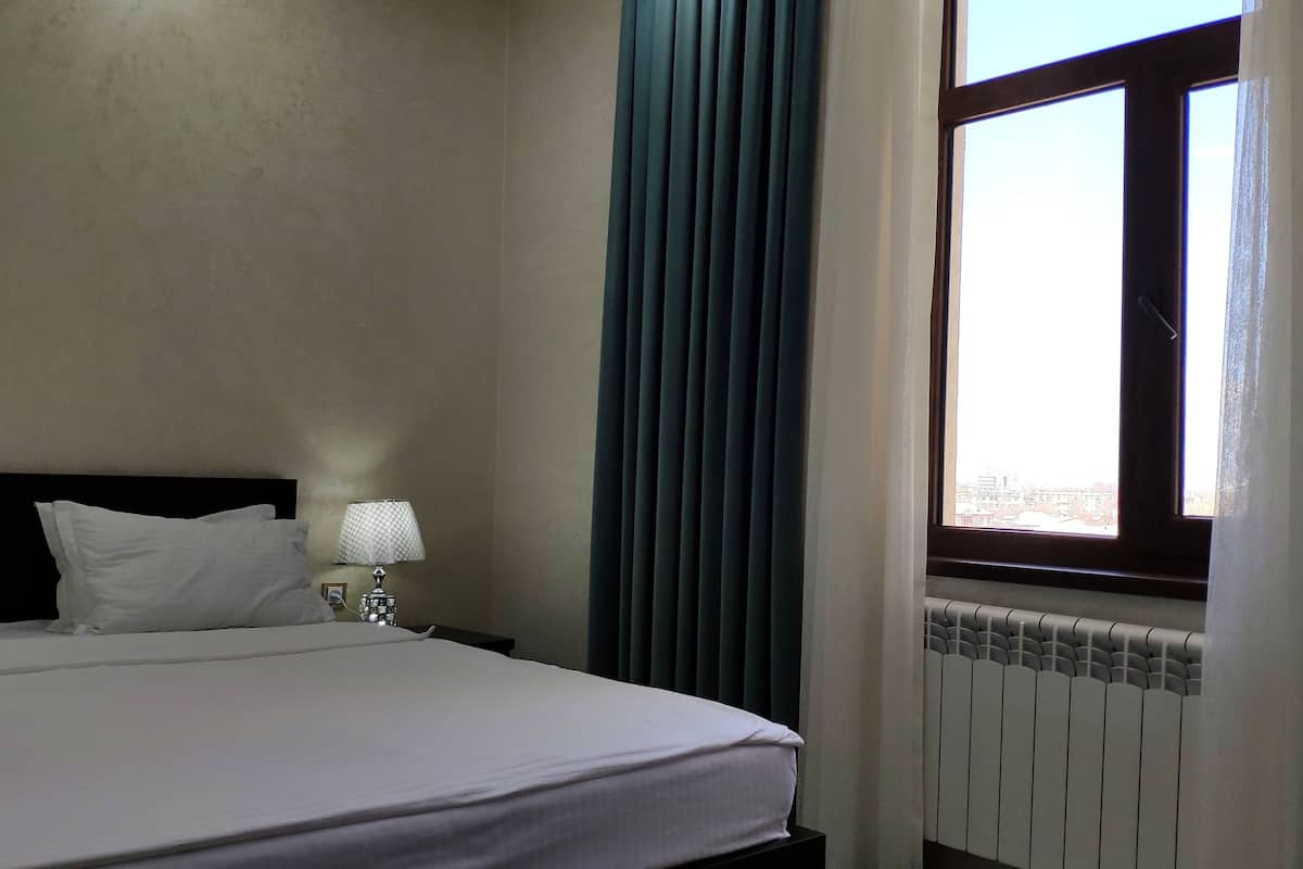 Ein Hotelzimmer mit Bett, Nachttisch mit Lampe, Fenster mit Vorhängen und Heizkörper.