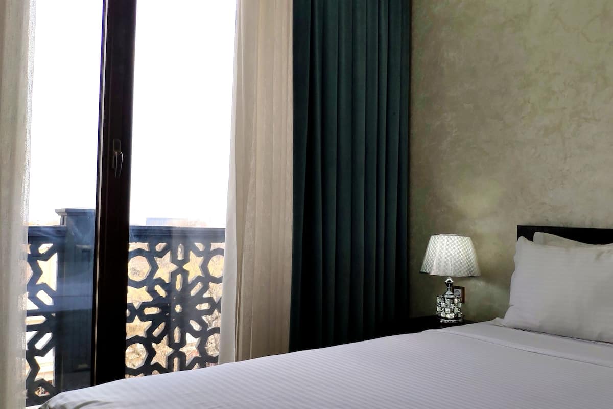 Ein Hotelzimmer mit Bett, Nachttisch mit Lampe und Blick durch ein mit Vorhängen versehenes Fenster nach draußen.
