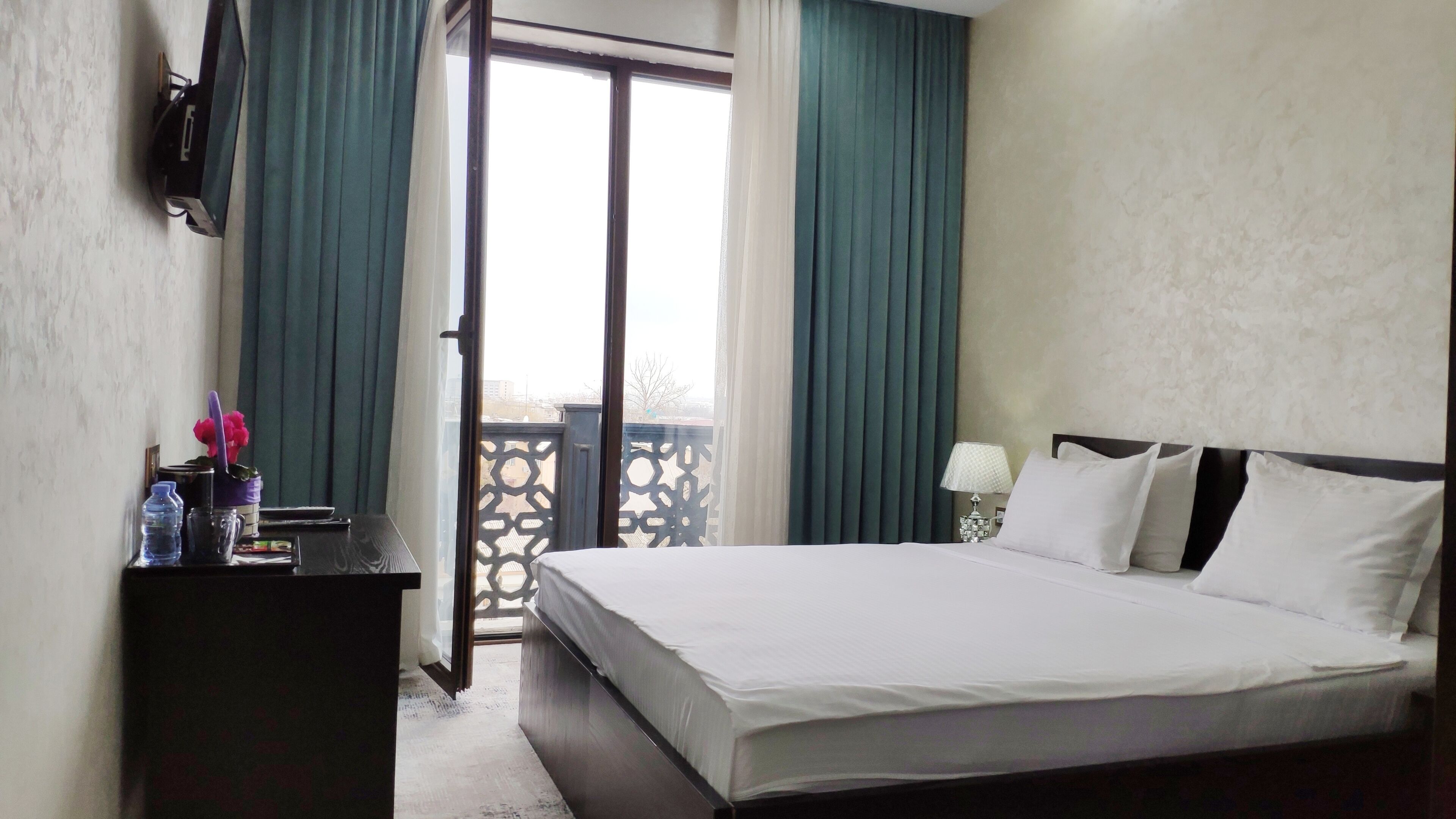 A hotel room with a bed, a desk with a lamp, a window with curtains, and a view of the outside.