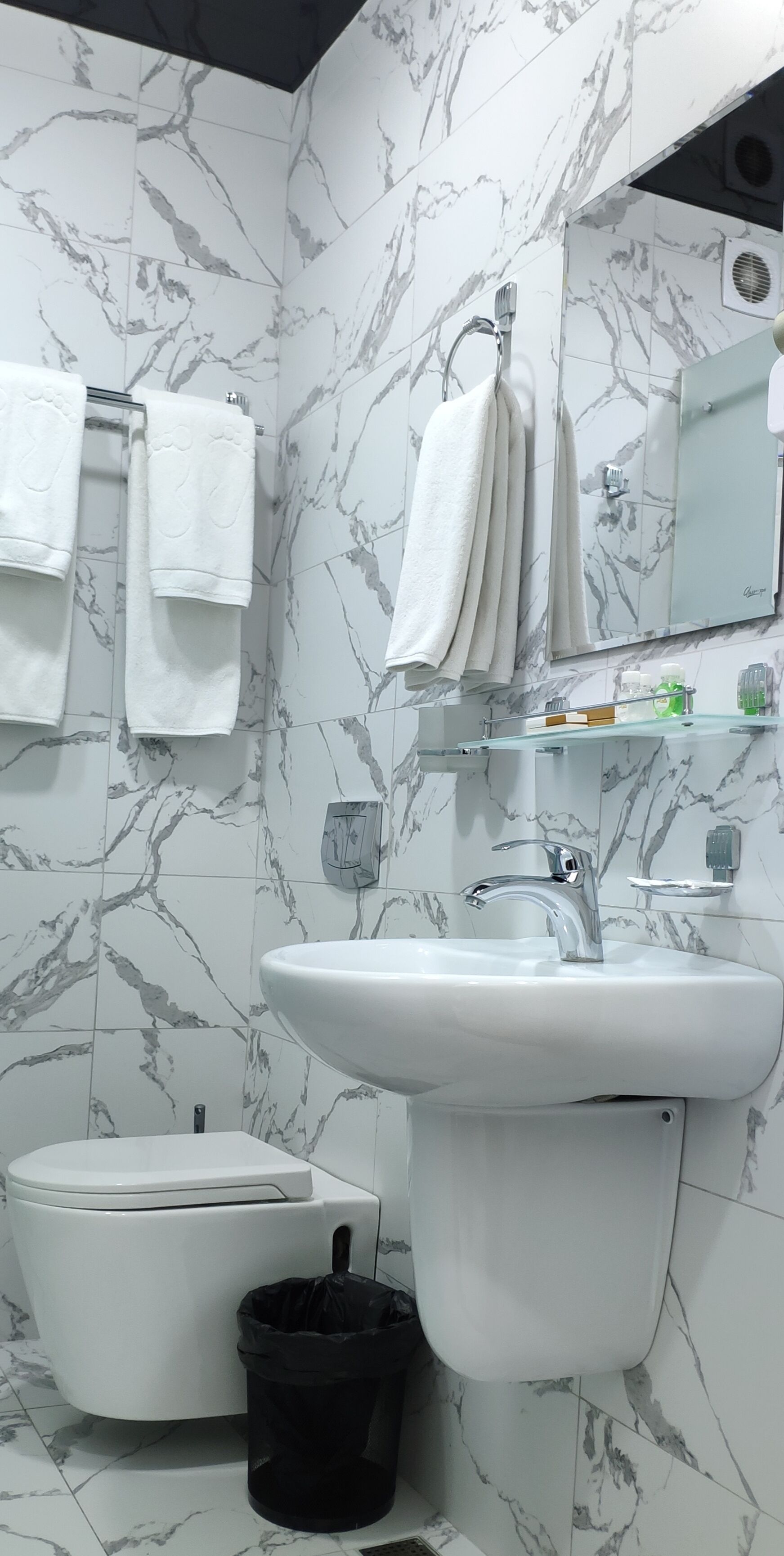 A bathroom with a white sink, a toilet, and marble-patterned walls. There are white towels hanging on a rack, and a black trash bin is visible.