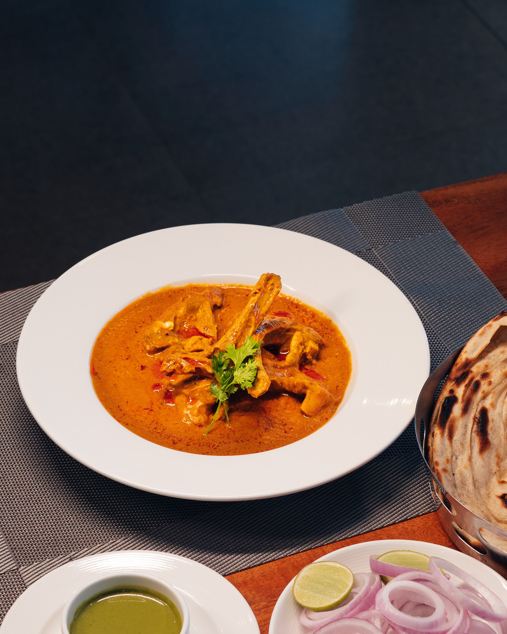 A plate of curry with chicken and a side of flatbread, accompanied by a small bowl of sauce and sliced onions.
