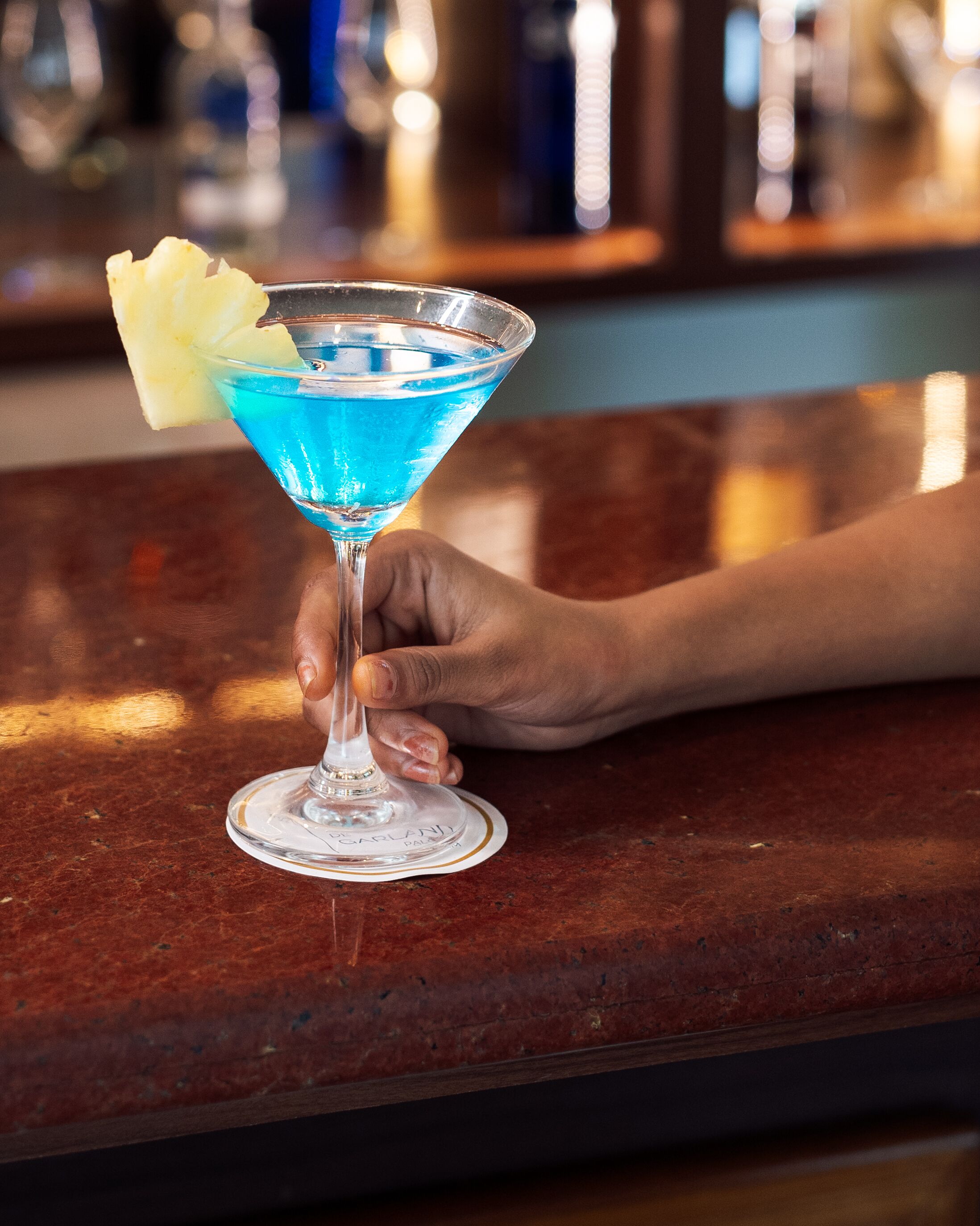 Una mano sostiene un cóctel azul con una rodaja de piña sobre una barra.