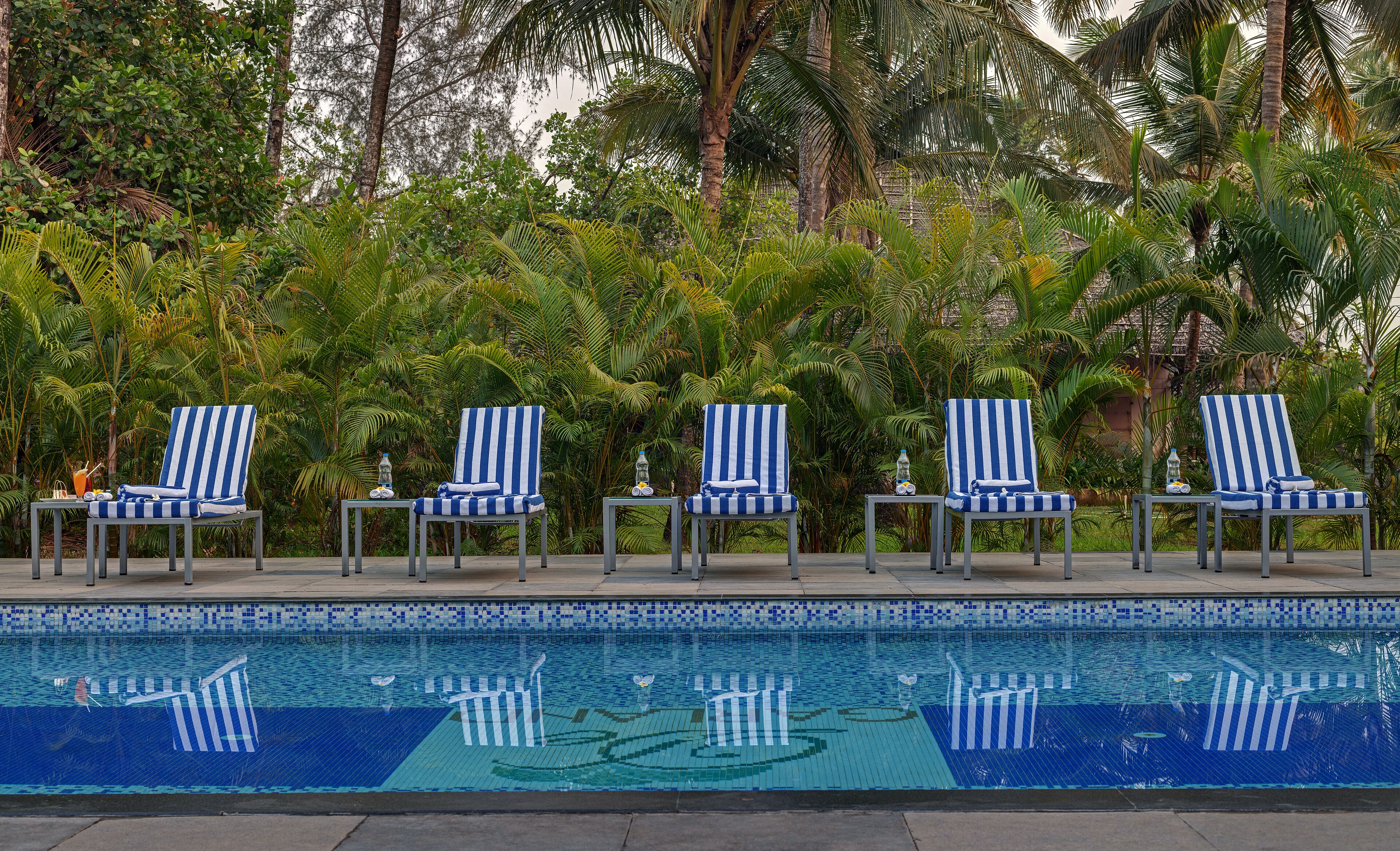 A poolside area with four striped lounge chairs, a tiled pool with a blue and white pattern, and lush tropical foliage in the background.
