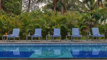 A poolside area with four striped lounge chairs, a tiled pool with a blue and white pattern, and lush tropical foliage in the background.