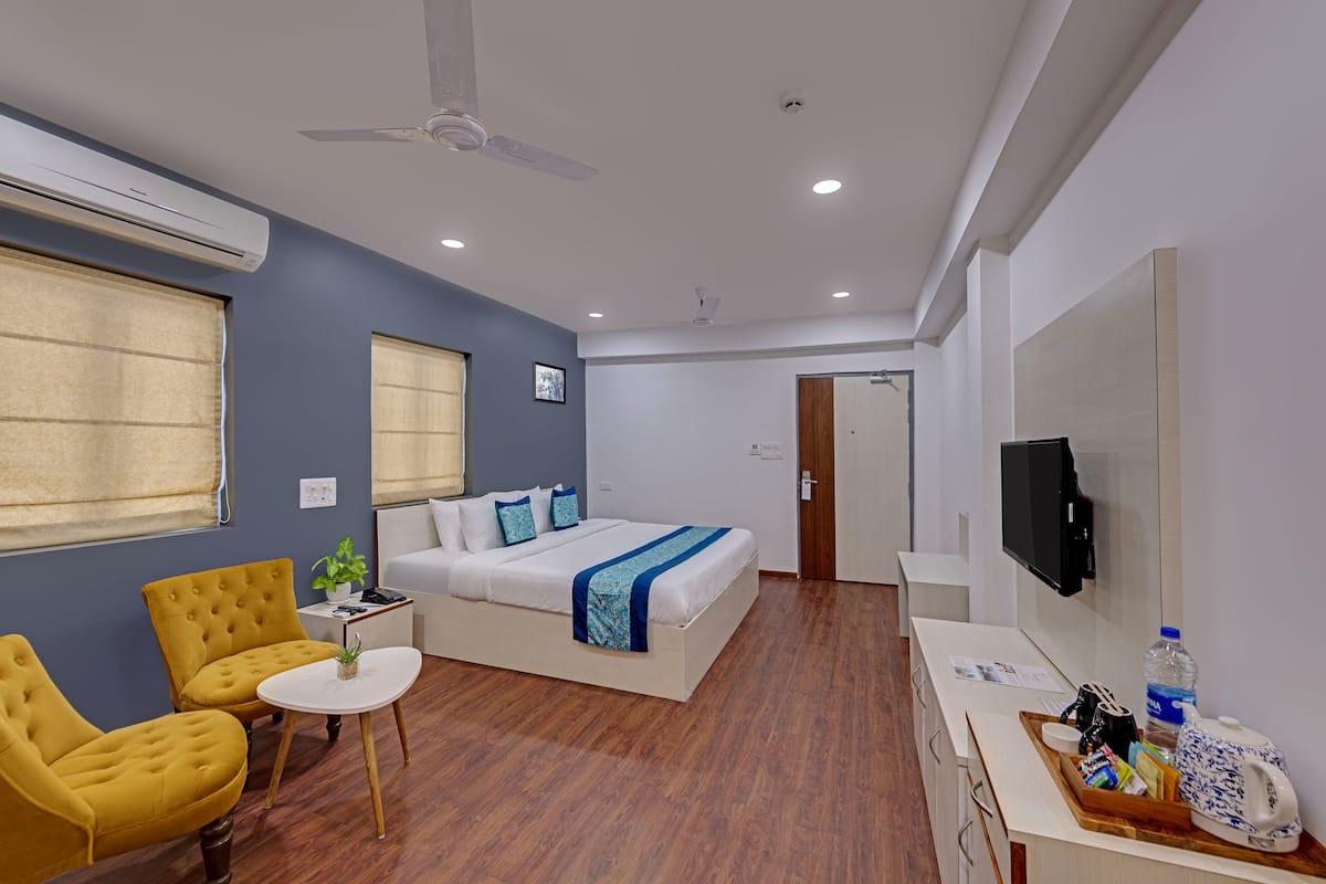 Une chambre d’hôtel avec un lit, un canapé, deux fauteuils, une petite table avec une plante, un téléviseur fixé au mur et un plancher en bois.
