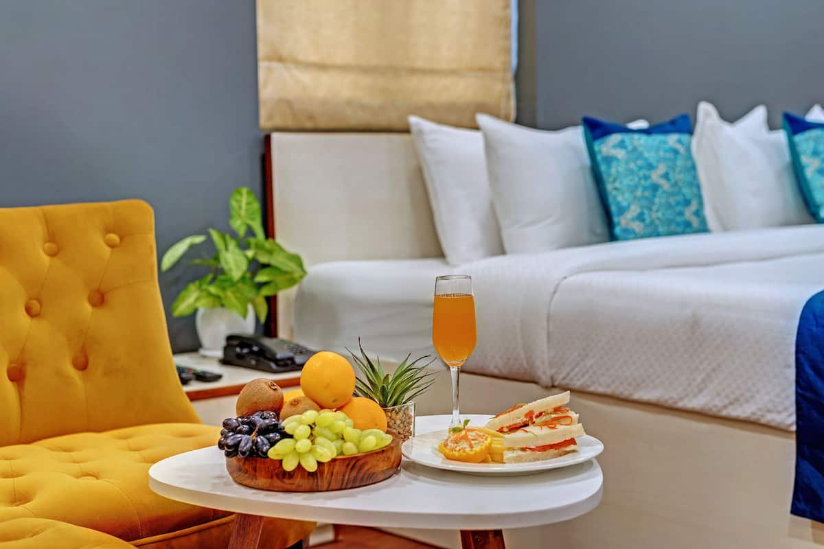 Une chambre d’hôtel avec un lit, une table de chevet sur laquelle se trouve un panier de fruits, un verre contenant une boisson et une chaise jaune.