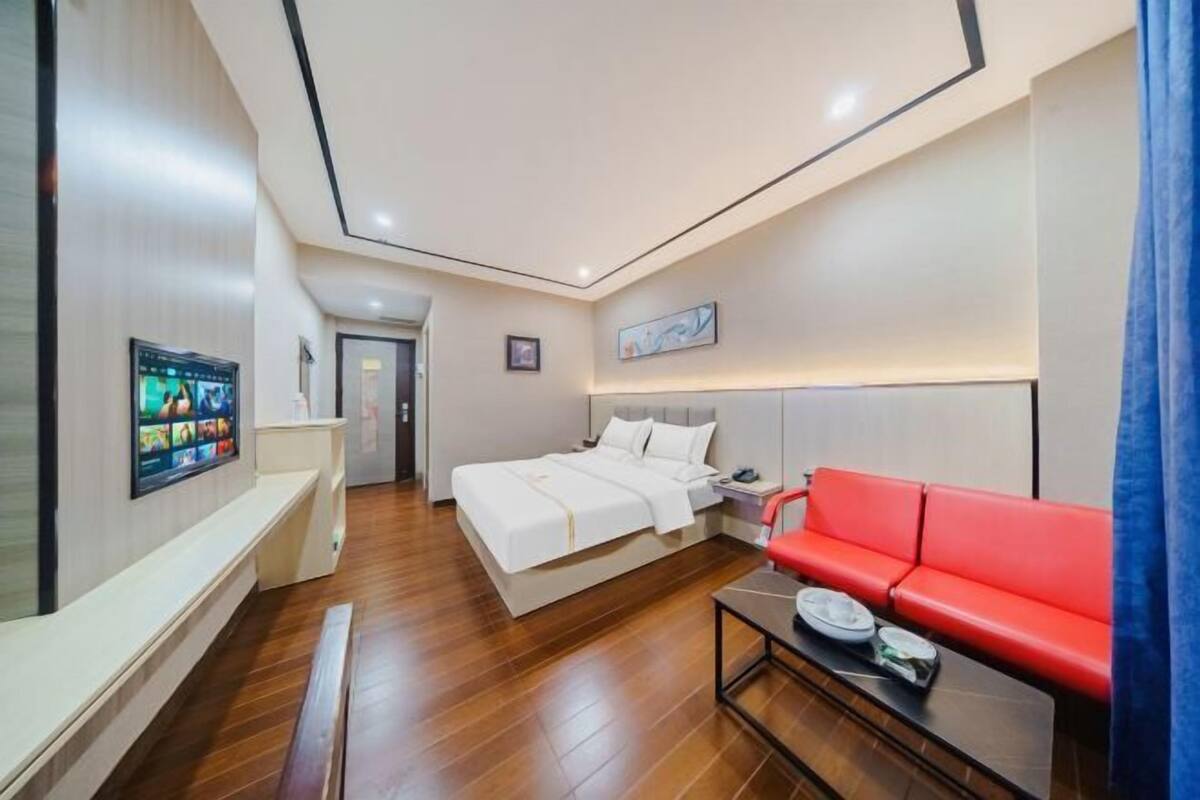 A modern hotel room with a bed, a red sofa, a TV, and a desk with a lamp.