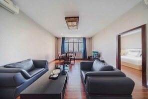 A modern hotel room with two black leather sofas, a dining table with chairs, a TV, and a large bed with a wooden headboard.