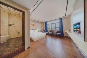 Una habitación de hotel moderna con una cama grande, una zona de estar con una mesa y sillas, un televisor montado en la pared y un baño con ducha e inodoro.