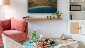 Un salón con un sofá rosa, una mesa redonda con aperitivos y bebidas, un cuadro de un paisaje costero y una estantería.