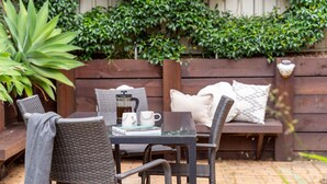 Un patio con una mesa de vidrio, sillas de mimbre, una cafetera y un banco con cojines.
