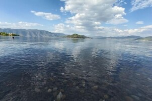 Um lago límpido com pedras visíveis e montanhas ao longe, sob um céu parcialmente nublado.
