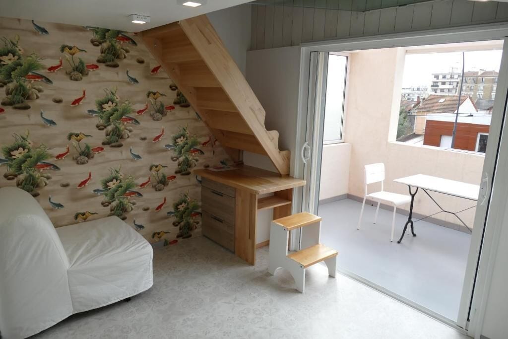 Una habitación con un escritorio de madera, una silla, una escalera y un balcón con vistas a edificios.
